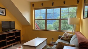 Ein Wohnzimmer mit einer Couch, einem Couchtisch, einem Fernseher auf einem Schrank, einem Fenster mit Blick ins Grüne und einer Lampe.