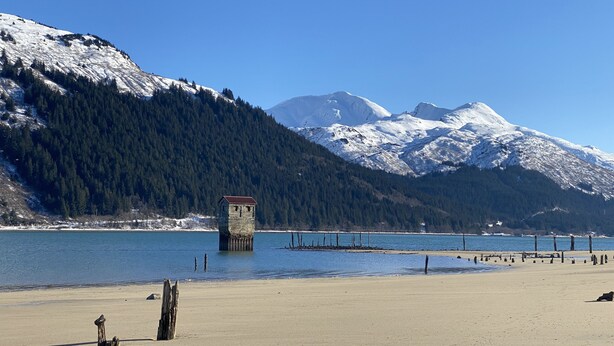 Ein Strand mit einer Holzkonstruktion im Vordergrund, bewölkter, klarer Himmel und schneebedeckte Berge im Hintergrund.