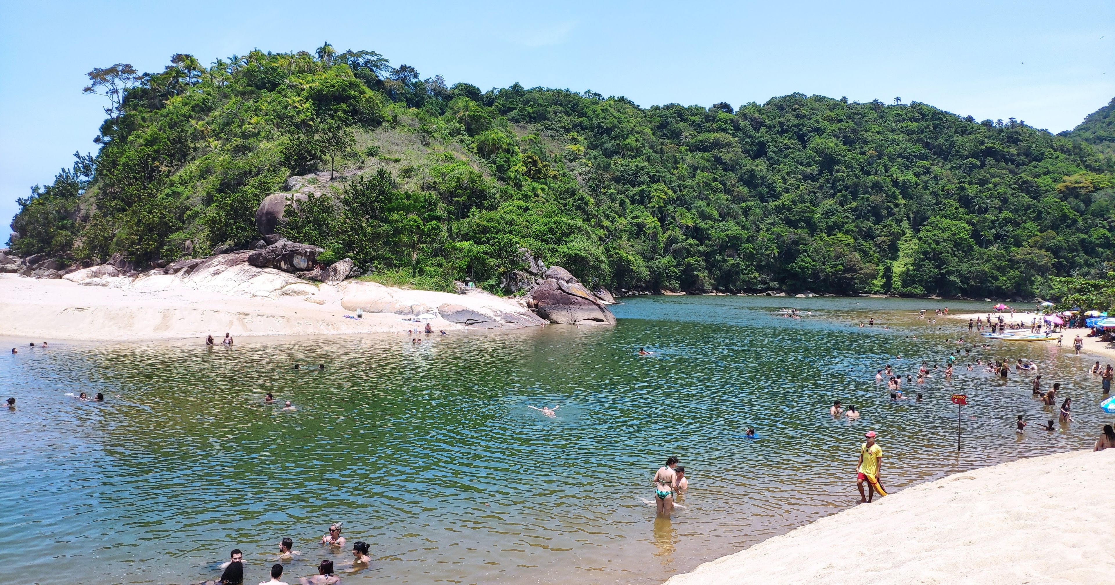 Uma praia com pessoas nadando e tomando sol, rodeada por vegetação exuberante e formações rochosas.