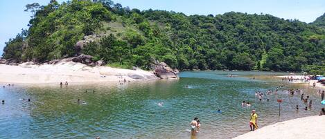 A beach with people swimming and sunbathing, surrounded by lush greenery and rocky outcrops.