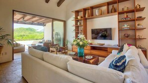 Amplia sala de estar con televisor grande, estantes de madera y vista a zonas verdes.