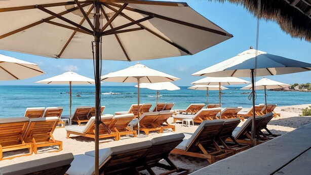 Un área de descanso frente a la playa, con sillones de madera y sombrillas grandes, con vista al océano.