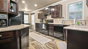 A modern kitchen with dark wood cabinets, a built-in oven, a coffee maker, and a window with blinds. - Newly Constructed, Single Level, Open Layout, Walk to Sandy River, 17 Miles to Government Camp (Rhododendron)