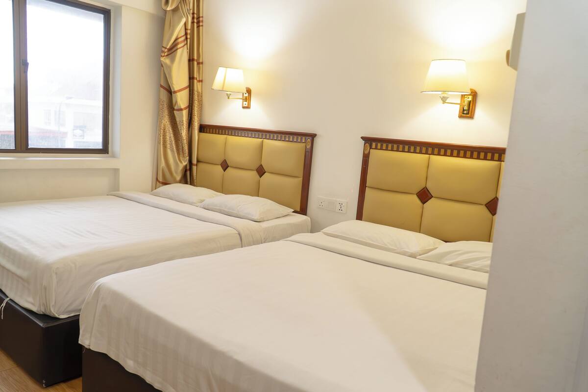 Une chambre d’hôtel avec deux lits, chacun doté d’une tête de lit, d’une fenêtre avec des rideaux et de lampes fixées au mur.