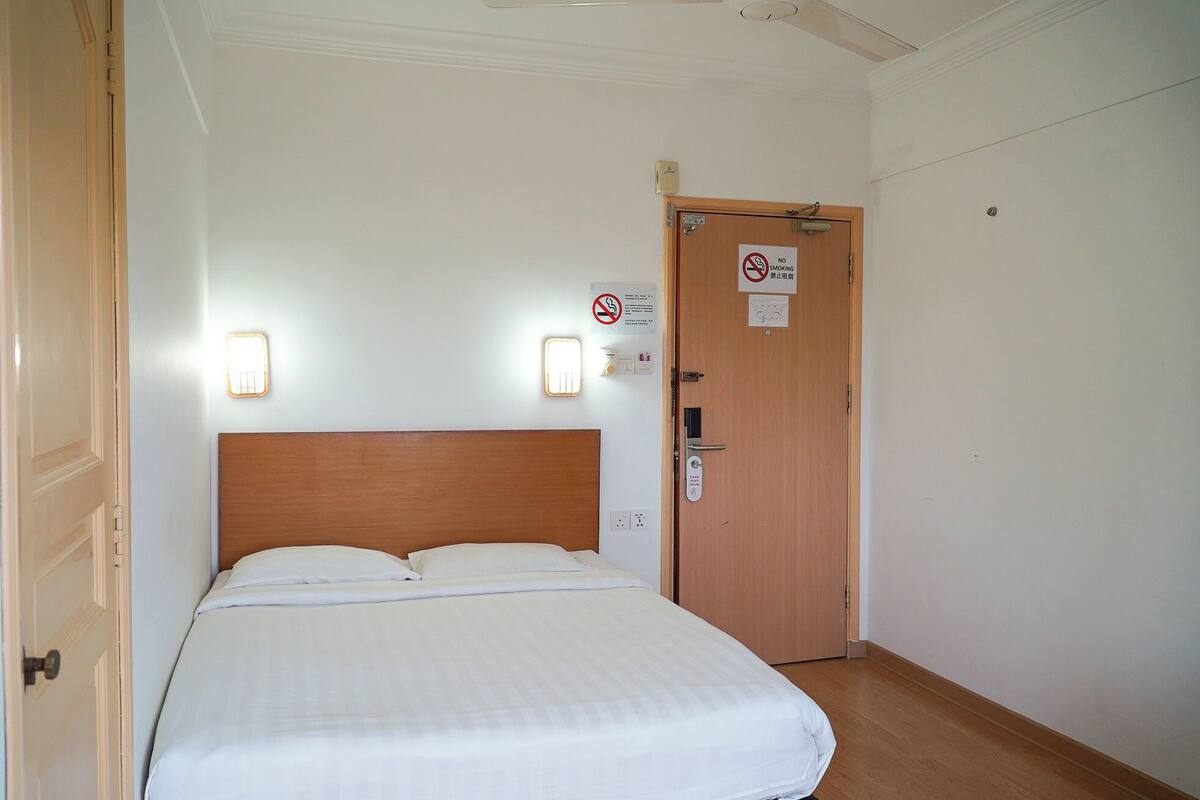 Une chambre avec une tête de lit en bois, un lit avec une literie blanche, une porte sur laquelle est apposé un panneau « interdiction de fumer » et un luminaire fixé au mur.