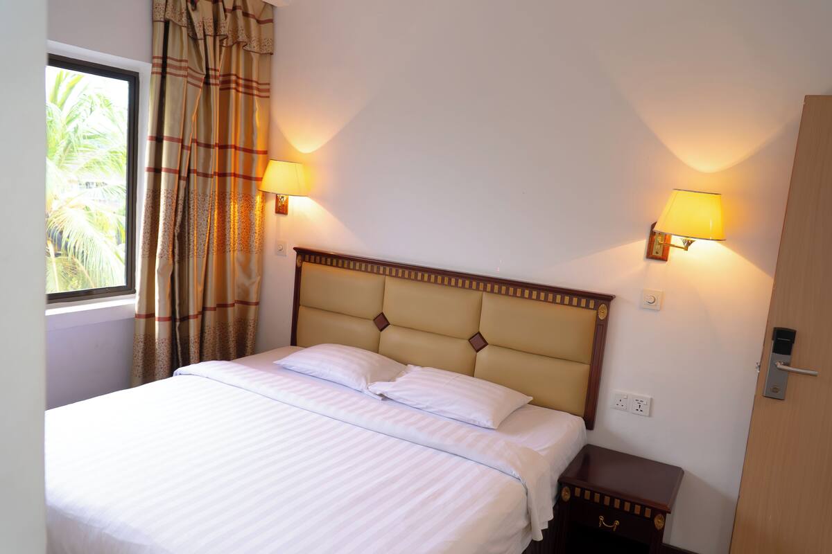 Une chambre d’hôtel avec un lit, deux lampes fixées au mur, une fenêtre avec des rideaux et une table de chevet.