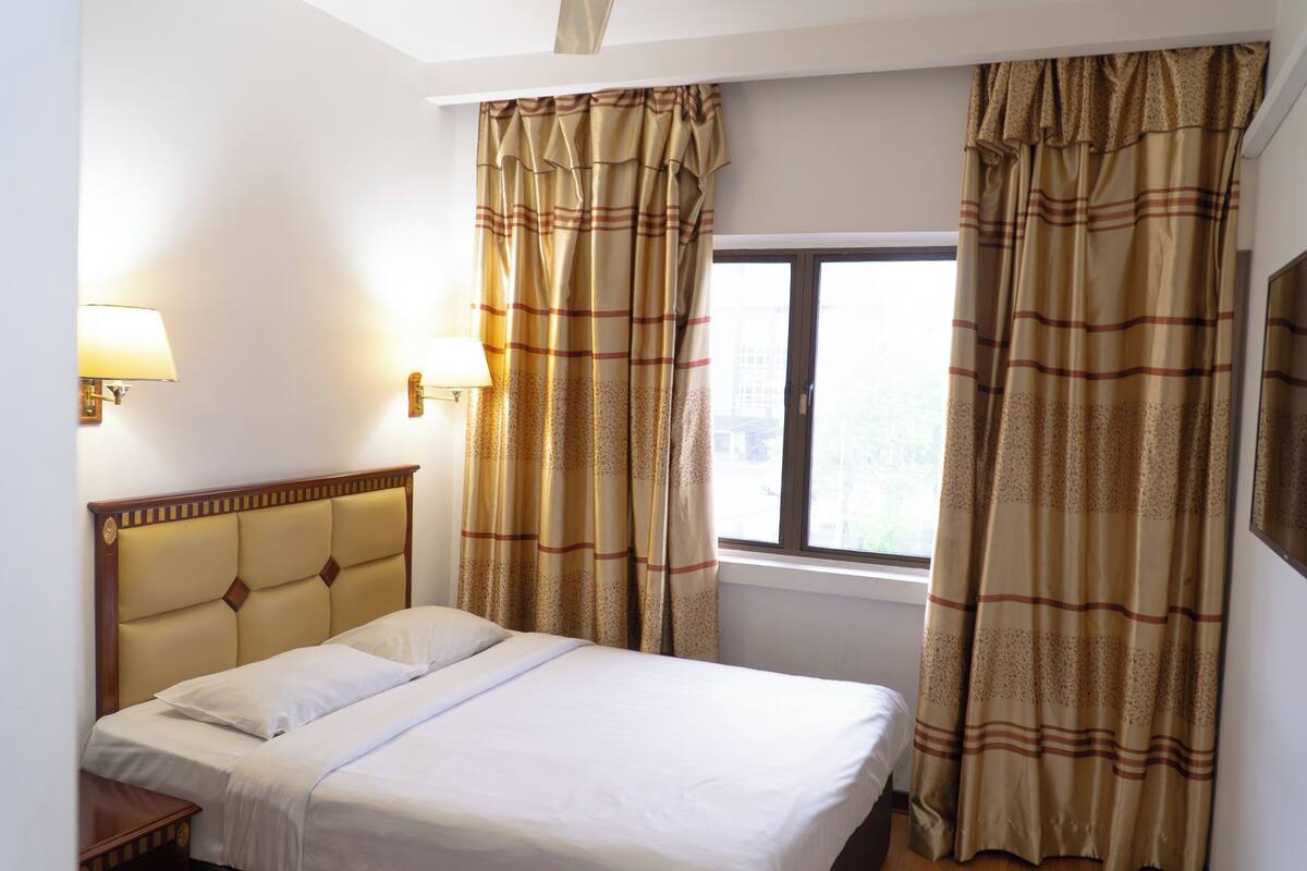 Une chambre d’hôtel avec un lit, deux lampes de chevet, une fenêtre et des rideaux.