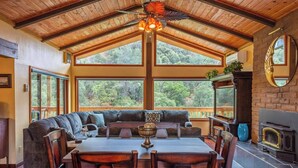 A cozy room with a wooden ceiling, large windows offering a view of the outdoors, a fireplace, and a dining area with a table and chairs.