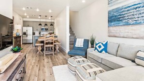 Un vaste espace de vie comprenant un canapé, des poufs et un tableau. On aperçoit à l’arrière-plan une cuisine équipée d’appareils modernes et dotée de meubles en bois.