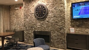 A hotel lobby with a stone wall, a large clock, a fireplace, a TV displaying a tennis match, and a seating area with a table and chairs.