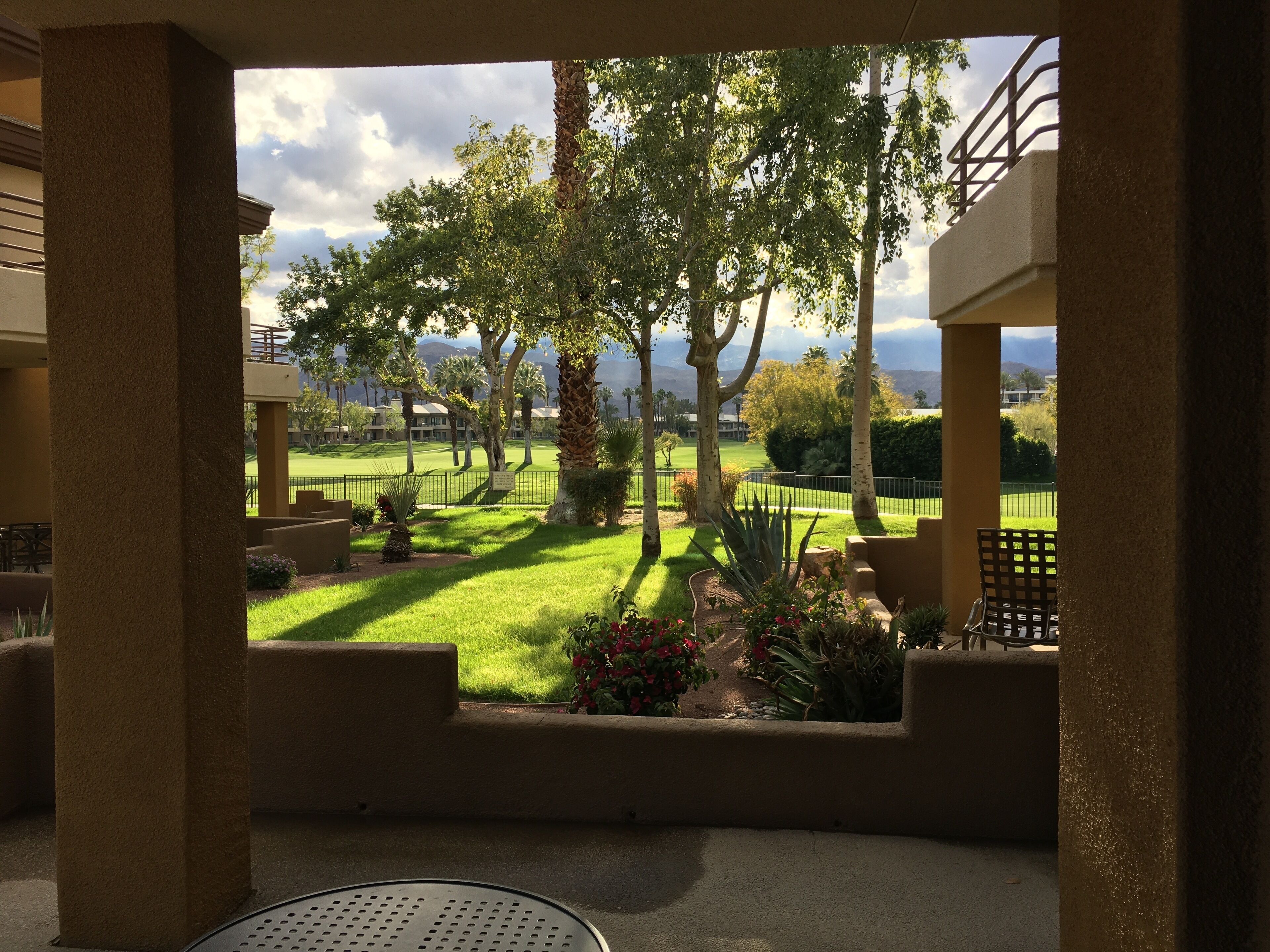 Vista de uma varanda com área de pátio, incluindo uma mesa e cadeiras, com vista para um gramado bem cuidado e árvores.