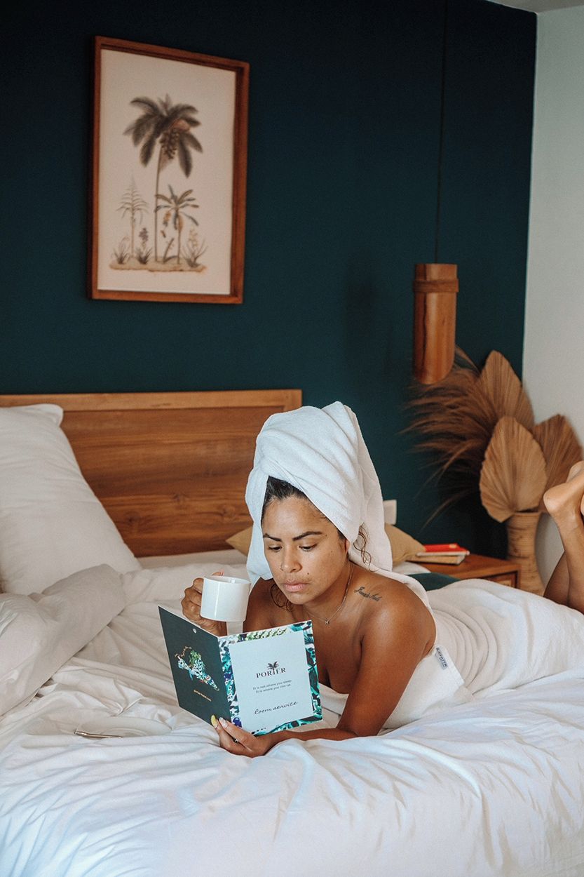 Uma pessoa deitada na cama, com um livro e uma xícara, em um quarto com cabeceira de madeira e um quadro na parede.