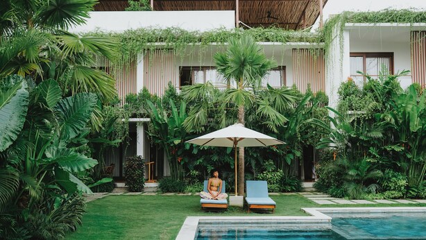 Un área junto a la piscina con una persona meditando, rodeada de exuberante follaje y un edificio moderno con grandes ventanales.