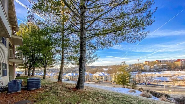 A residential area with a large tree in the foreground, a building with a balcony on the left, and a view of a community with multiple houses and a parking lot in the background. - Branson Condo on Thousand Hills Golf Course with Indoor Pool and Hot Tub (Branson)
