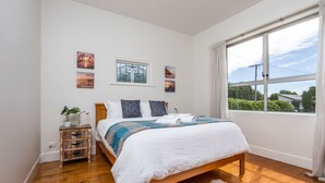 Ein Schlafzimmer mit einem hölzernen Bett, weißer Bettwäsche, einem kleinen Nachttisch mit Pflanze und einem Fenster mit Blick auf Grünflächen.