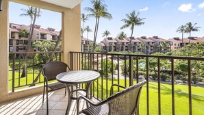 A balcony with a table and chairs, overlooking a pool and palm trees, with buildings in the background.