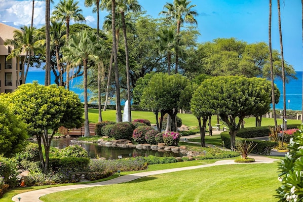 A lush garden with palm trees, a pond, and a pathway leading to a building with a view of the ocean.
