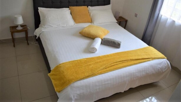 A neatly made bed with white and yellow bedding, a black headboard, and a small bedside table with a lamp. - North Manor Nakuru (Nakuru)