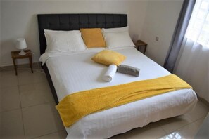 Una cama bien tendida con ropa de cama blanca y amarilla, un cabecero negro y una mesita de noche con una lámpara.