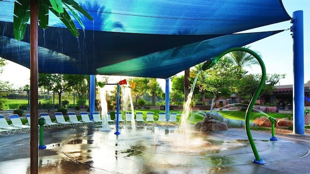 A water play area with multiple water jets, a shaded seating area with white lounge chairs, and a surrounding landscaped garden. - Marriott's Shadow Ridge - Enclaves - 2 Bedroom (Palm Desert)