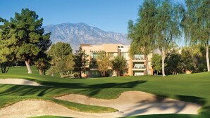 A golf course with bunkers and trees, a building with multiple floors and large windows, and mountains in the background. - Marriott's Shadow Ridge - Enclaves - 2 Bedroom (Palm Desert)