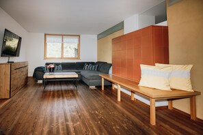 A modern living room with a wooden floor, a grey sectional sofa, a wooden bench, a TV on a wooden cabinet, and a window with a view.