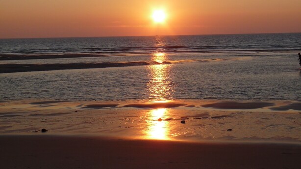 Ein Sonnenuntergang über einem Strand mit ruhigem Wasser und einer Person, die am Ufer entlangspaziert.