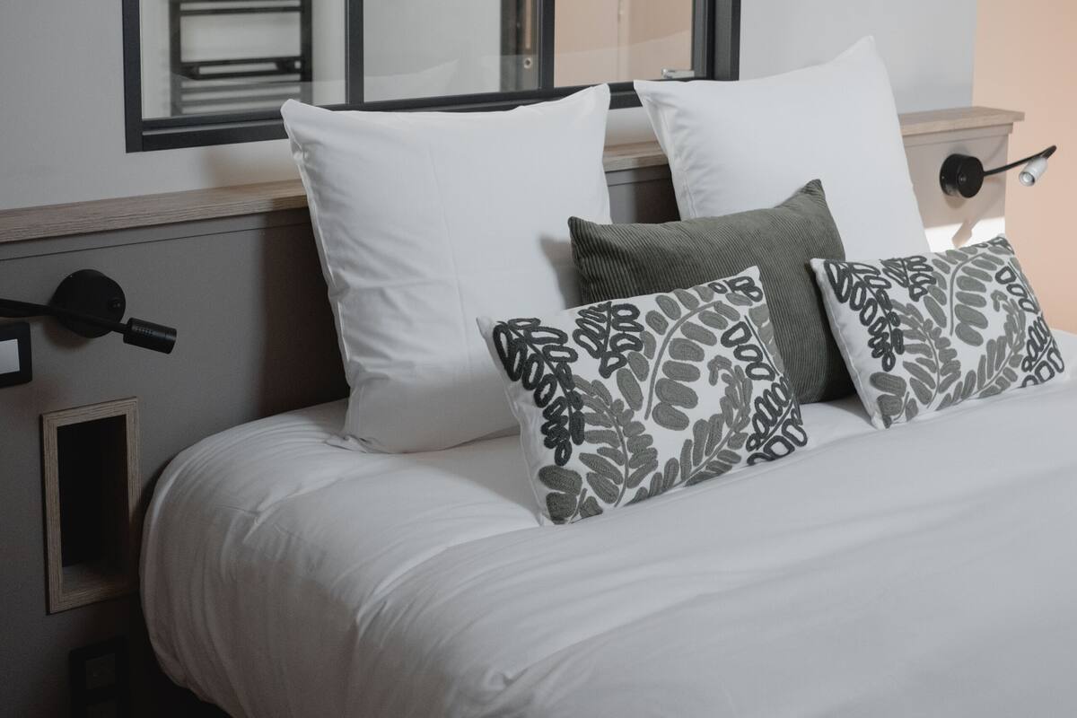 A neatly made bed with white bedding and patterned pillows, a large mirror above it, and a bedside table with a lamp.
