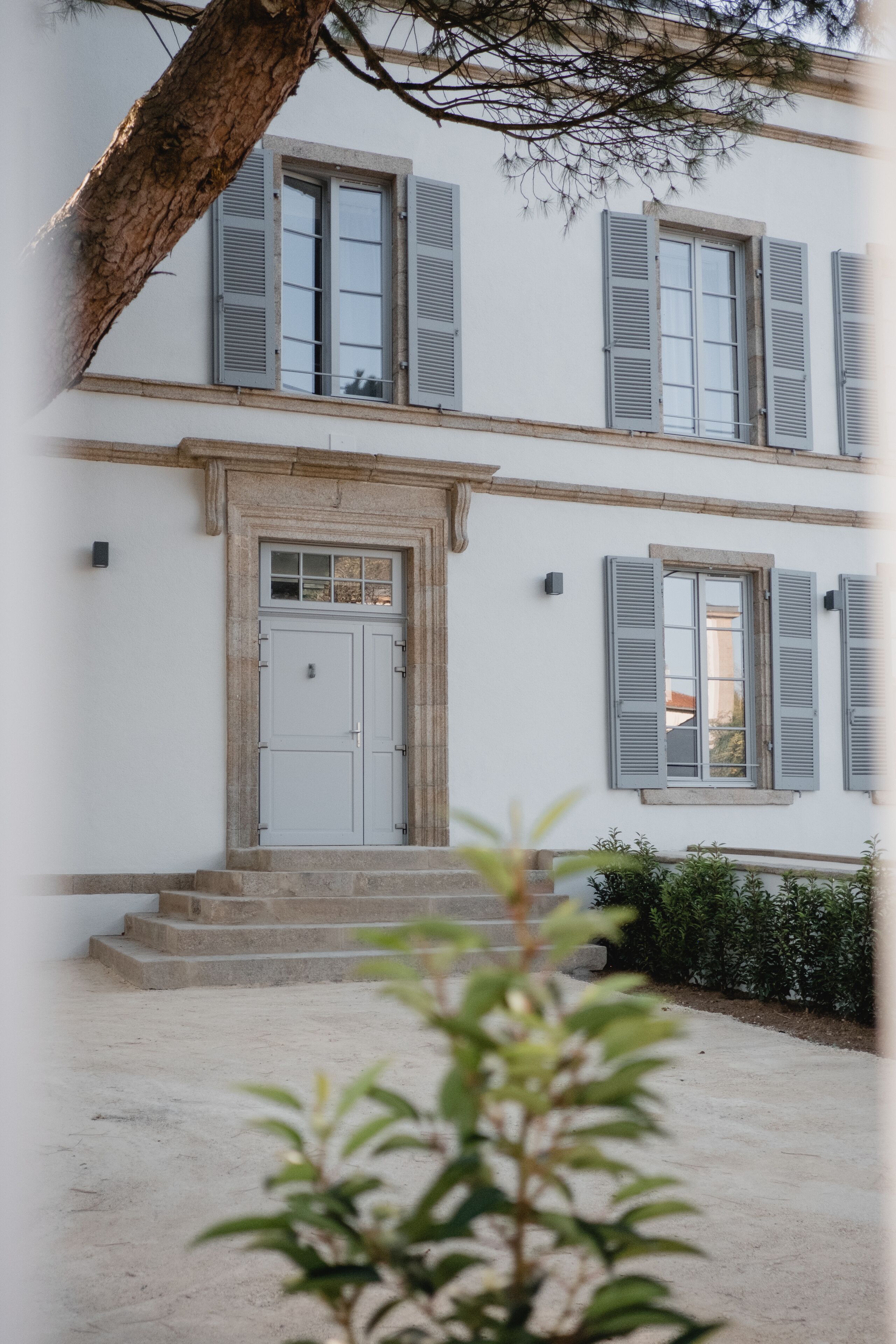 A two-story building with a white facade, grey shutters, and a central entrance with steps.