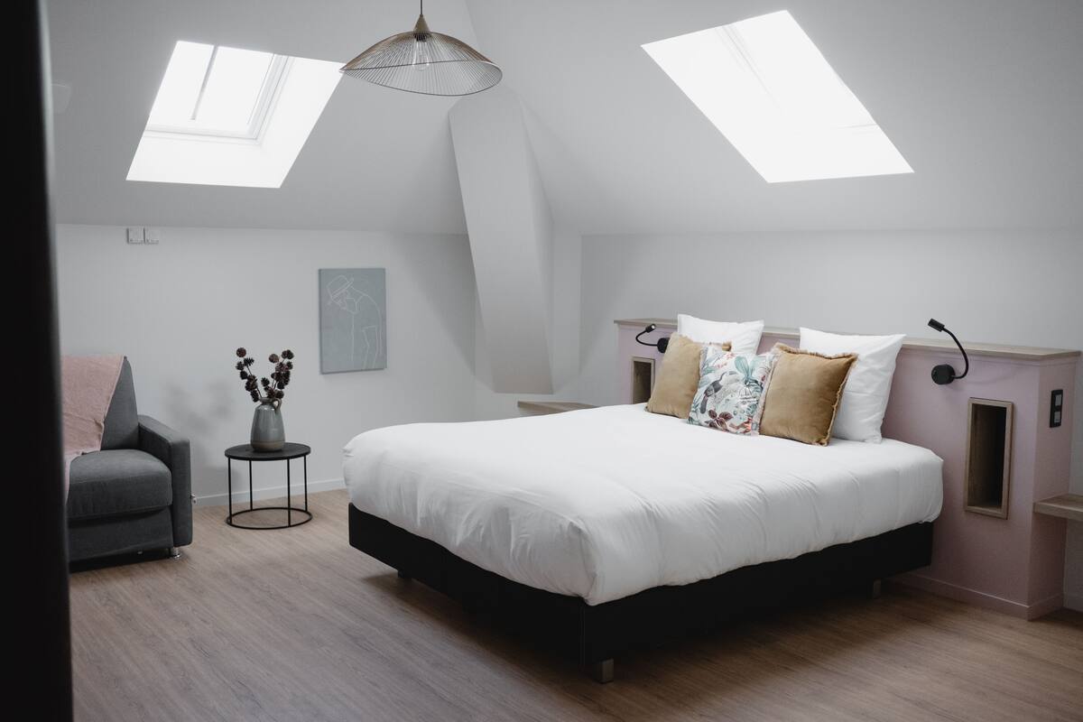 A modern bedroom with a large bed, skylights, a gray armchair, and a small side table.