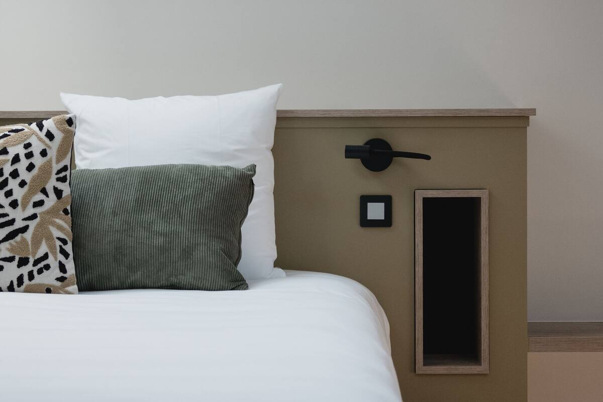 A neatly made bed with white and green pillows, a headboard with a light switch, and a small picture frame on the wall.