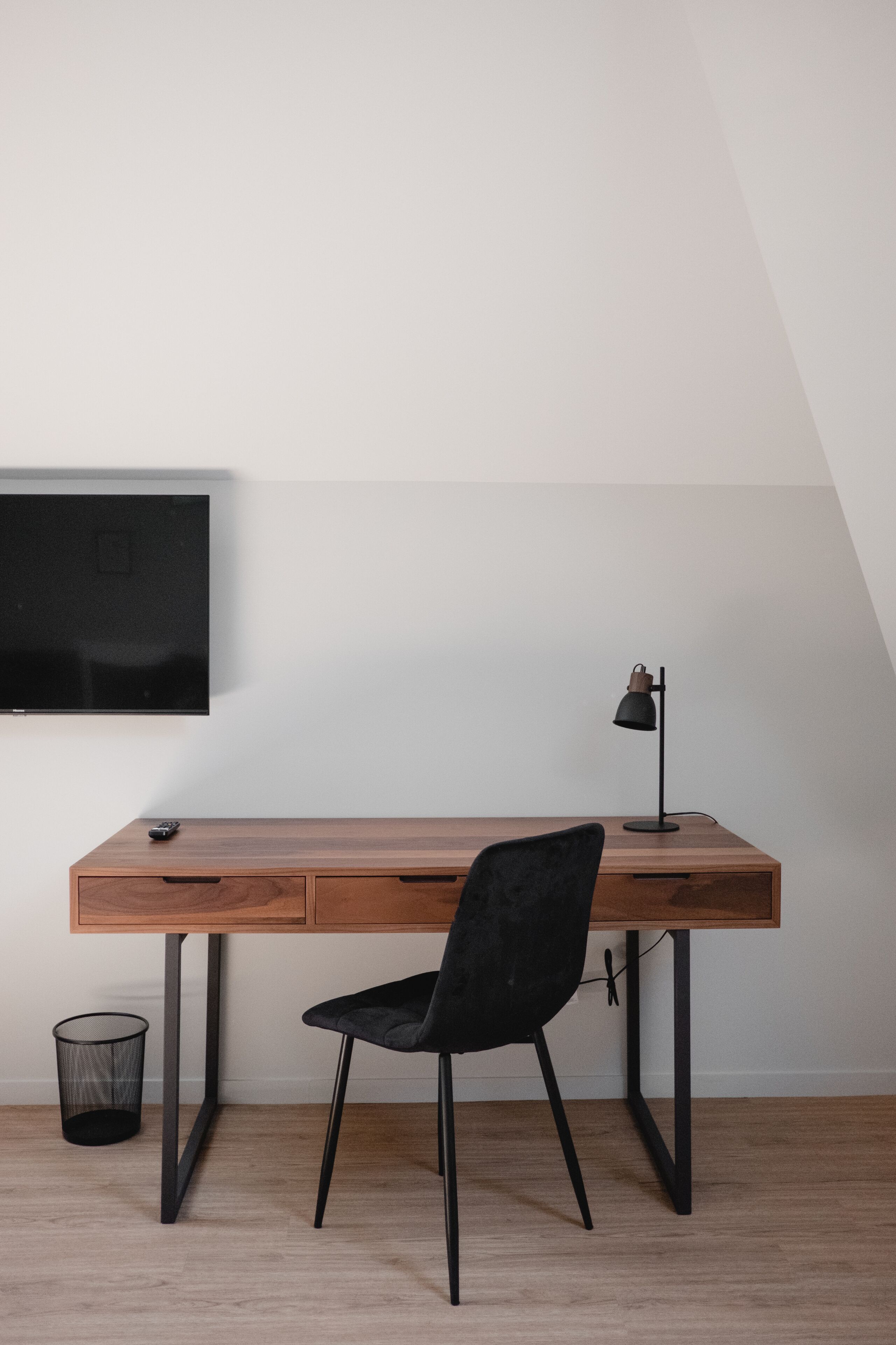 A wooden desk with a black chair, a television mounted on the wall, and a desk lamp.