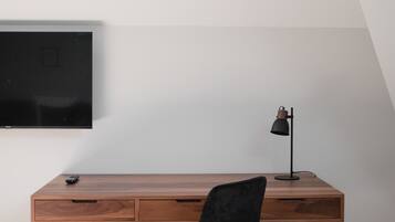 A wooden desk with a black chair, a television mounted on the wall, and a desk lamp.