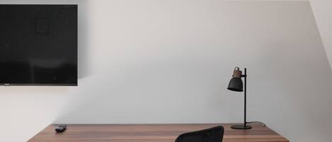 Un escritorio de madera con una silla negra, un televisor montado en la pared y una lámpara de escritorio.