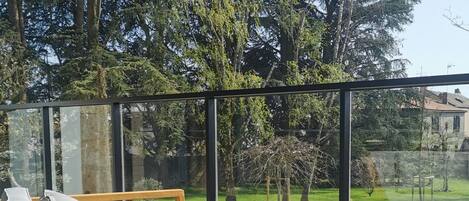 A wooden sofa with cushions, a small table with a teapot and cups, and a glass railing overlooking a garden with trees and a lawn.