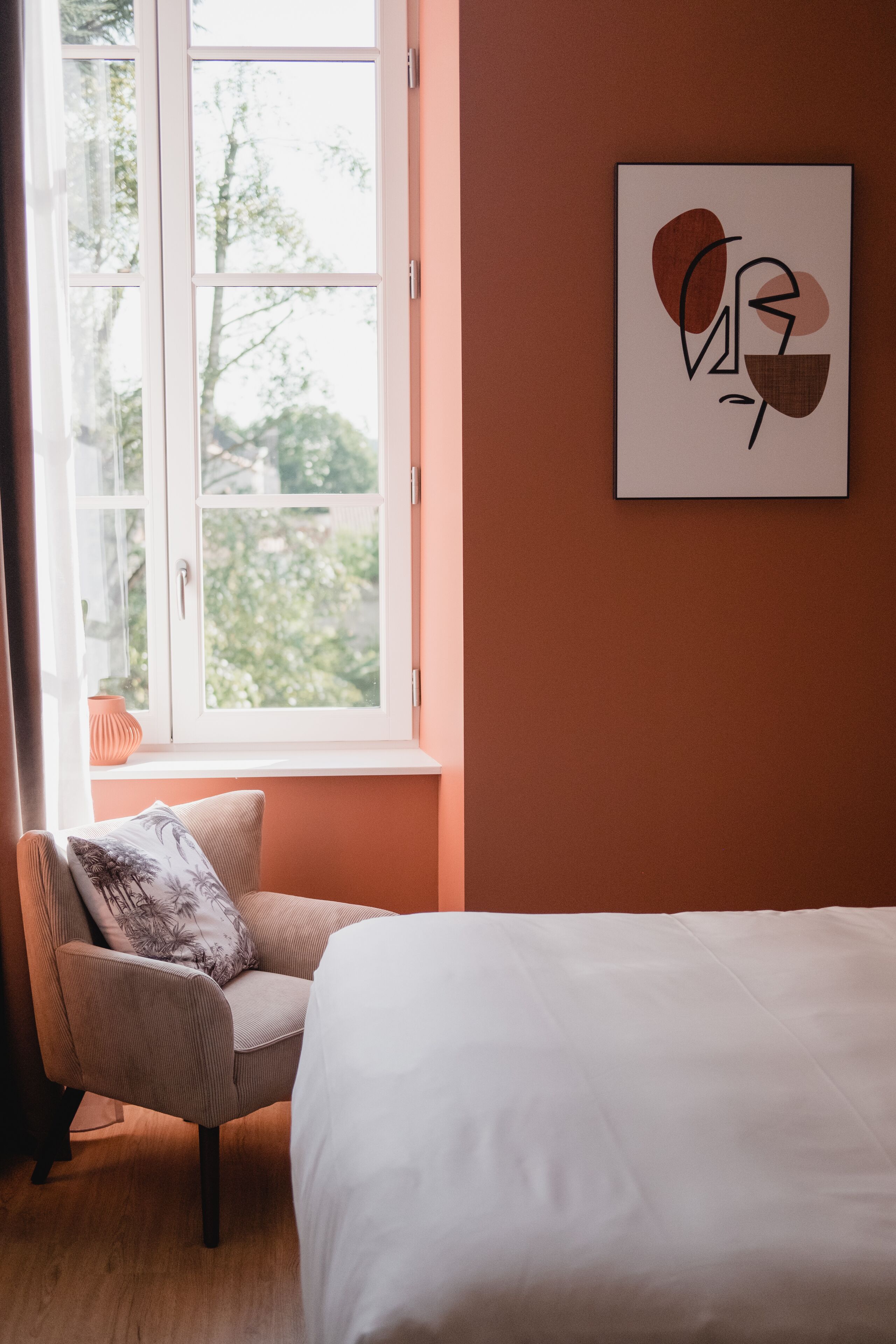 A bedroom with a bed, a chair with a pillow, a window, and a wall-mounted abstract art piece.