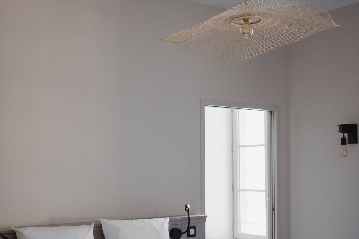 A bedroom with a bed, white bedding, two brown pillows, a hanging light fixture, a window, and a door.
