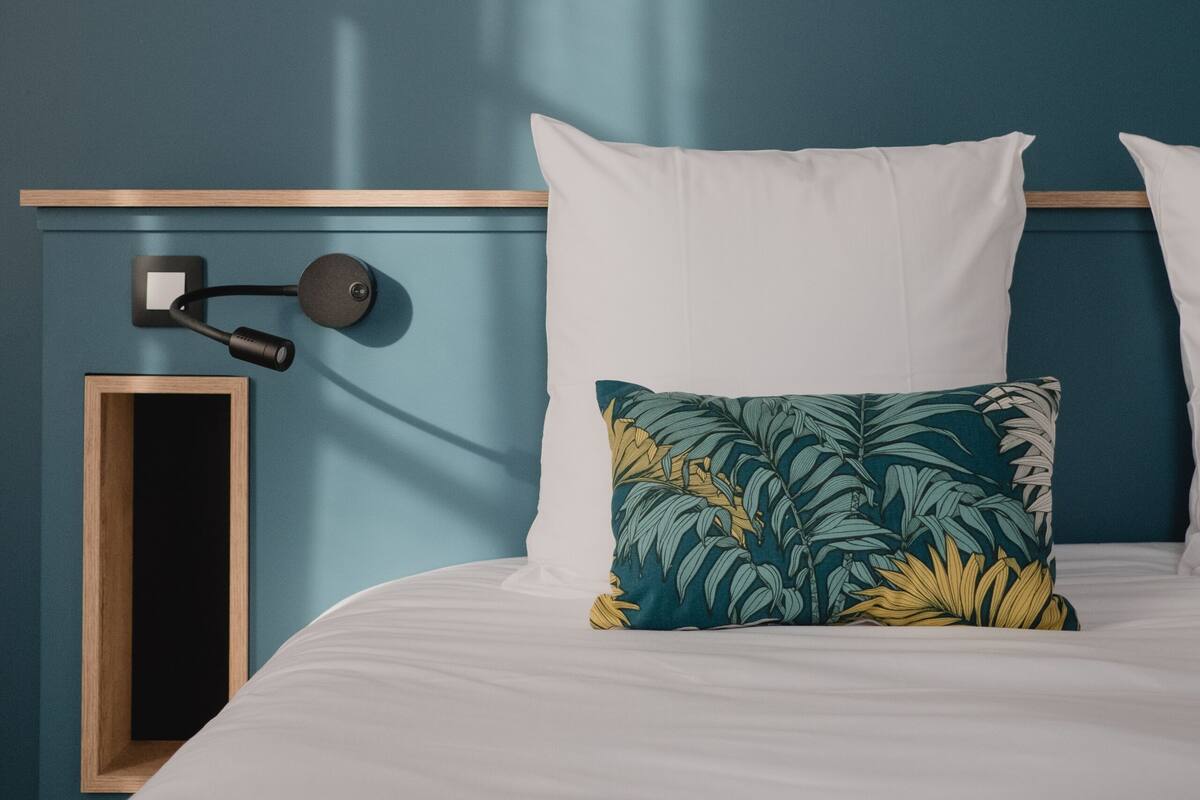 A neatly made bed with white and tropical leaf-patterned pillows, a framed botanical print on the wall, and a bedside lamp.