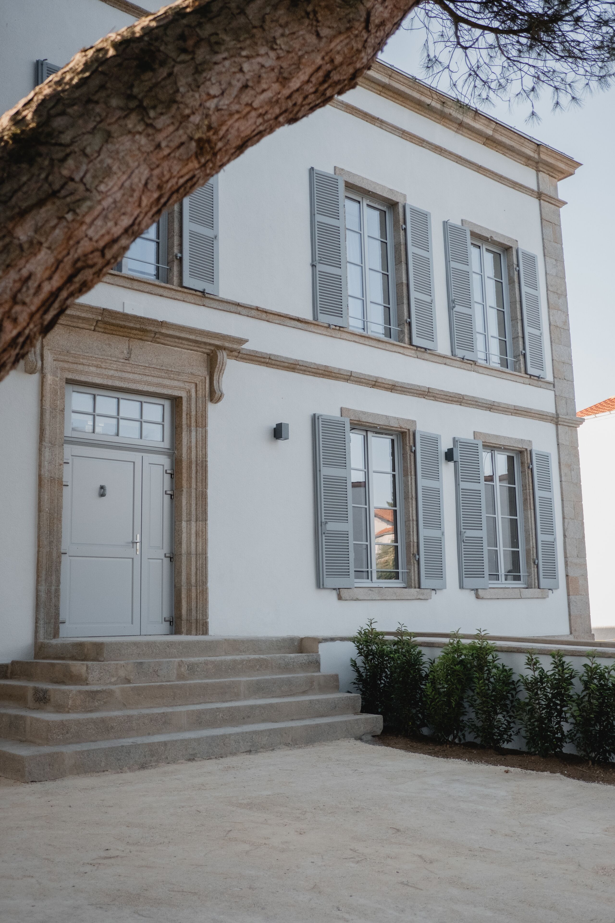 Un edificio de dos pisos con fachada blanca, contraventanas grises y una puerta central con un pequeño ventanal encima.