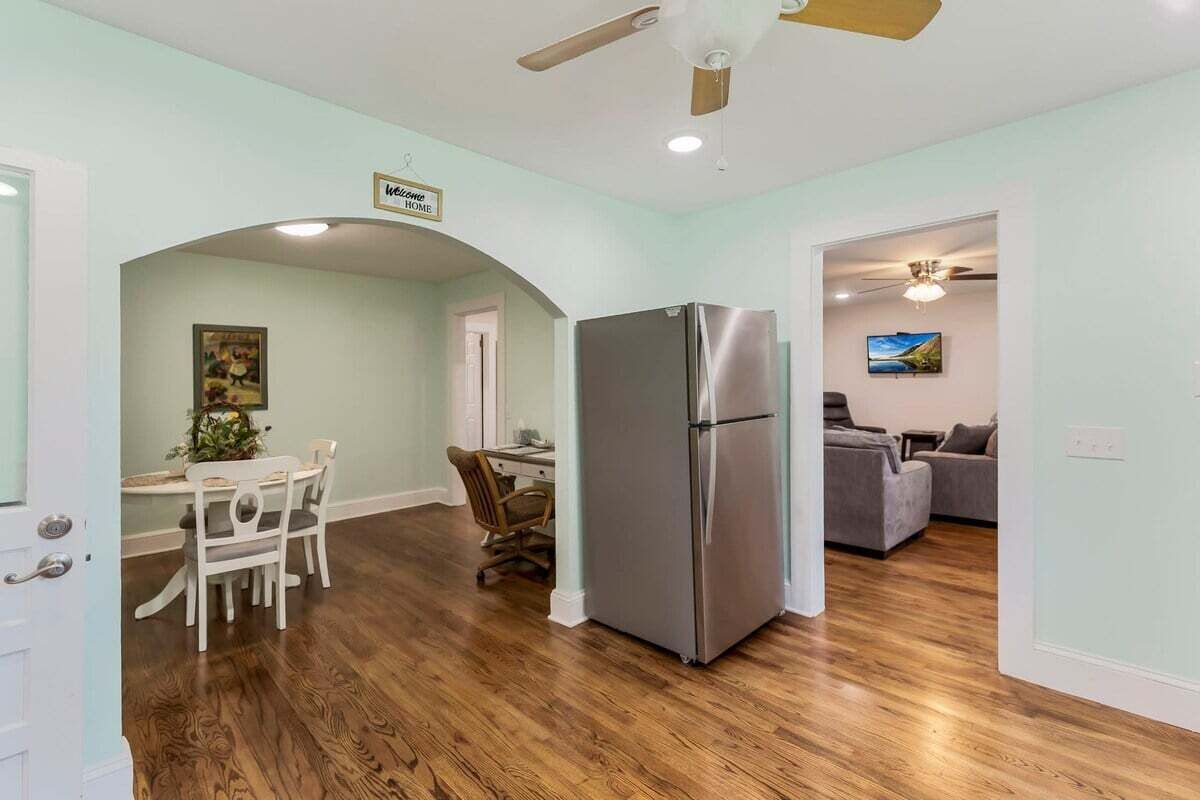 A spacious living area with hardwood flooring, a large stainless steel refrigerator, a dining table with chairs, and a sofa in the background.