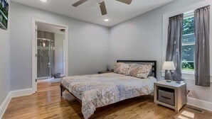 A bedroom with a bed, a nightstand, a ceiling fan, a window with curtains, and a door leading to a bathroom.