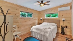 Une pièce de type cabane en bois, comprenant un lit, deux fenêtres, un ventilateur de plafond et un petit radiateur.