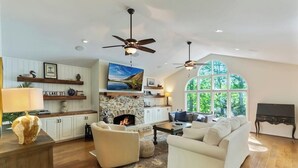 A spacious living room with a fireplace, white sofas, a coffee table, and a large window with a view of greenery.
