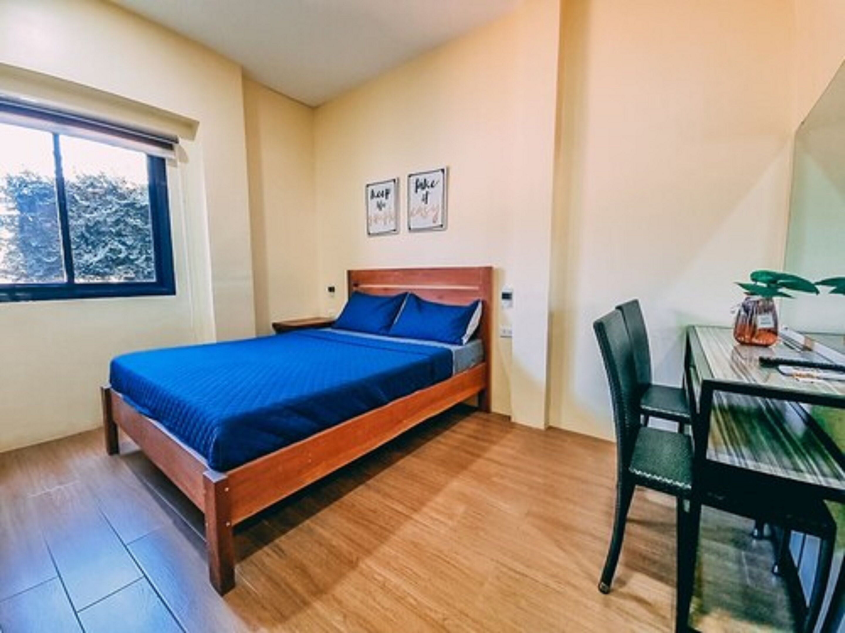 A small, neatly arranged bedroom with a wooden bed, a small table with a chair, and a window with a view of trees.