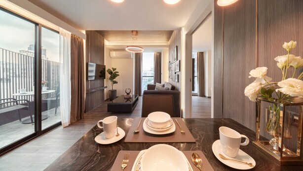 A modern dining area with a dark marble table set for four, large windows offering a city view, and a cozy seating area in the background. - Aspira Parc Sukhumvit 22 (Bangkok)
