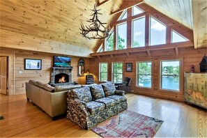A cozy cabin interior with a fireplace, a sofa, a patterned couch, and large windows overlooking a lake.
