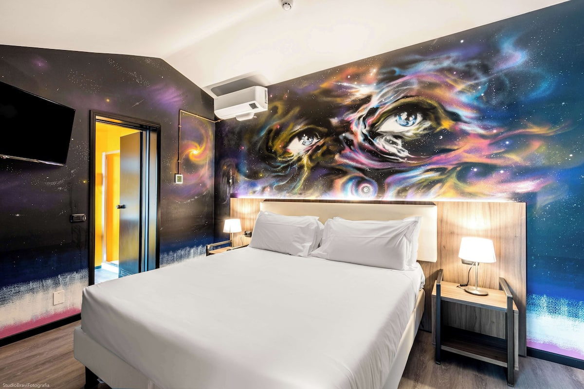 A hotel room with a large bed, a nightstand with a lamp, a wall-mounted TV, and a door leading to another room.