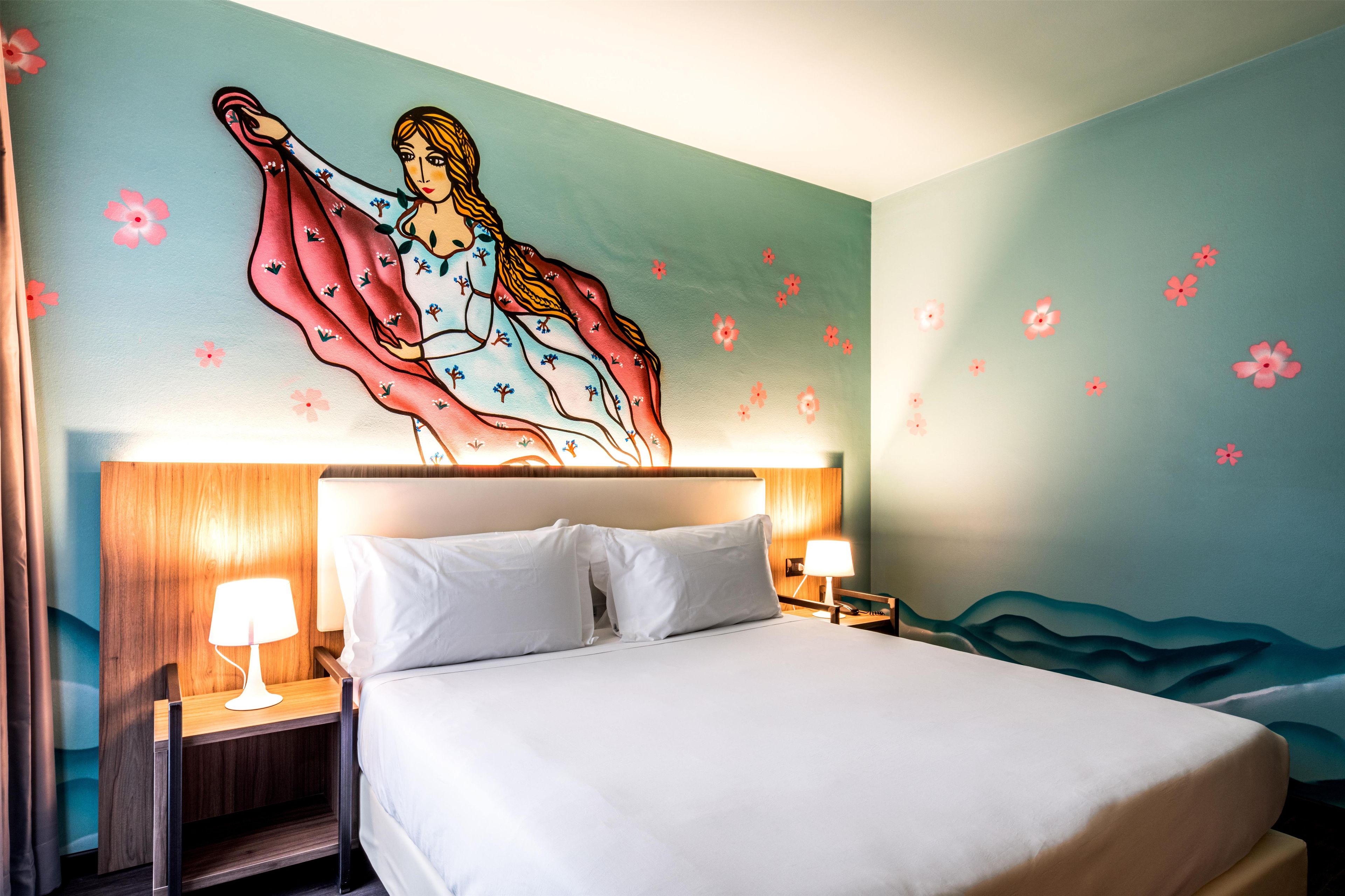 Una habitación de hotel con cama, dos lámparas de noche, un mural en la pared y una mesita de noche.