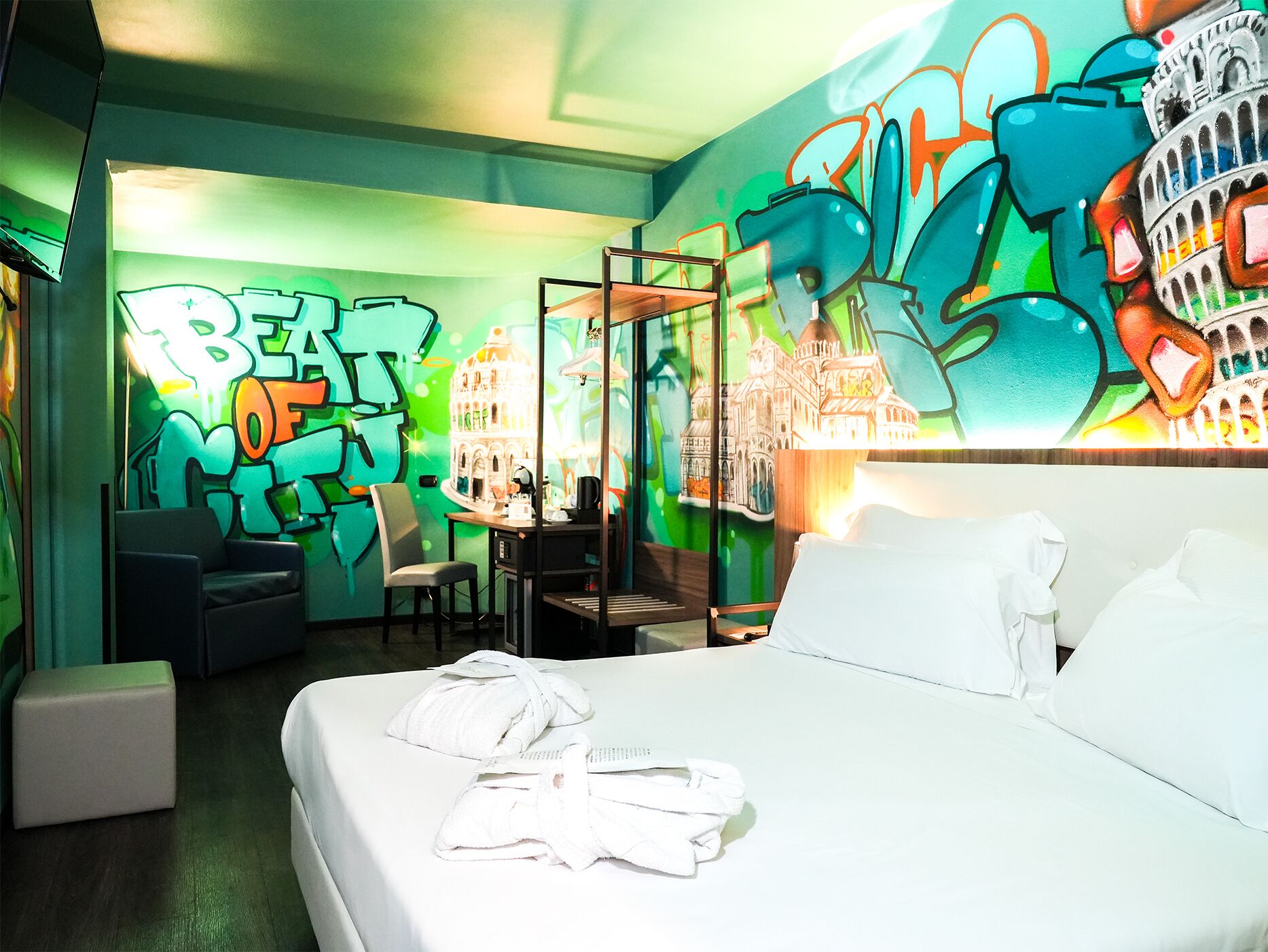 Habitación de hotel con cama, escritorio y sillas. La pared es verde y tiene arte callejero.