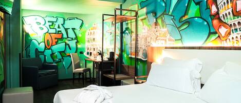 Habitación de hotel con cama, escritorio y sillas. La pared es verde y tiene arte callejero.
